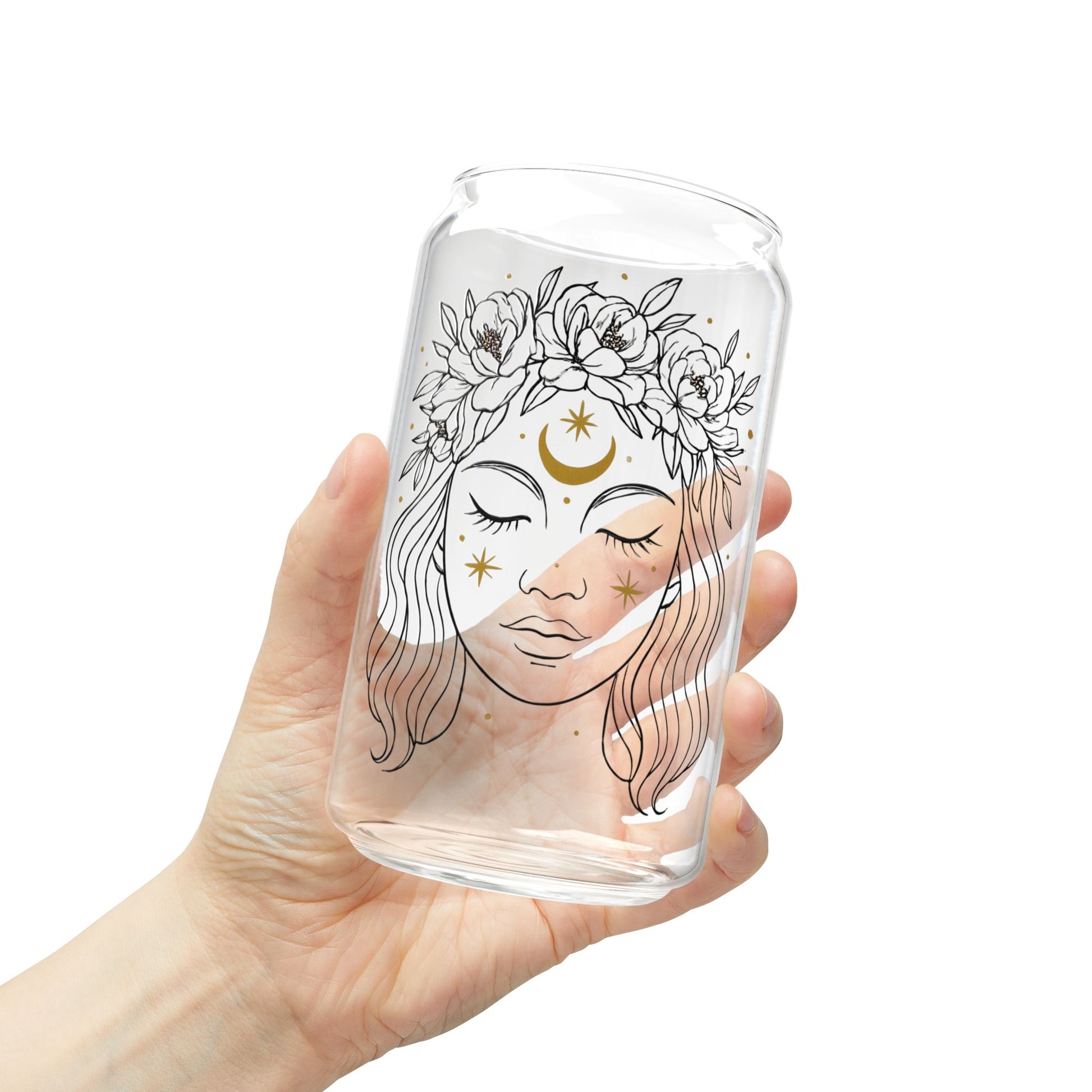 Clear glass tumbler with a decorative design of a face with flowers and a moon, held by a hand against a white background.