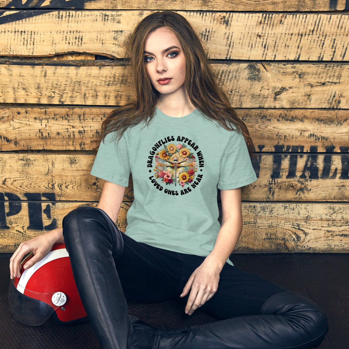 Woman wearing a dusty blue t-shirt with a graphic design, sitting against a wooden wall.